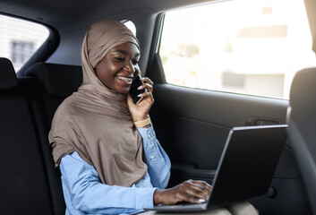 Smiling black islamic businesswoman talking on phone and using laptop in car