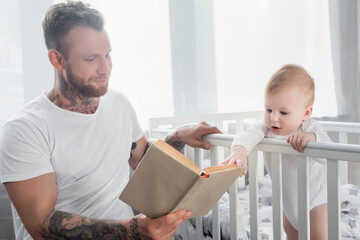 Fototapeta premium young tattooed father reading book to baby boy standing in cot