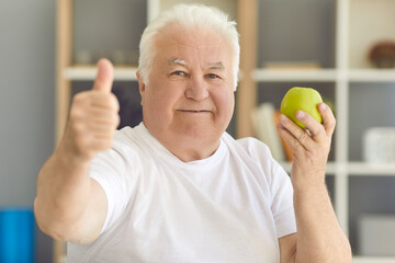 Happy senior man looking at camera holding fresh green apple and giving thumbs-up