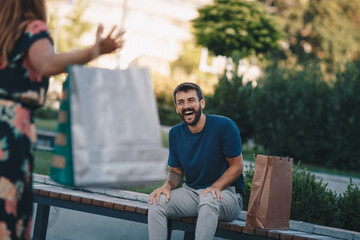 Fototapeta premium Smiling handsome young man sitting on the bench and waiting to his girlfriend. Girlfriend holding bags. Shopping concept.