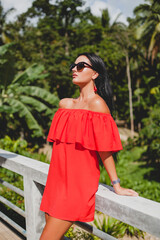 young stylish sexy woman in red summer dress standing on terrace in tropical hotel, palm trees background, long black hair, sunglasses, ethnic earrings, sunglasses, smiling