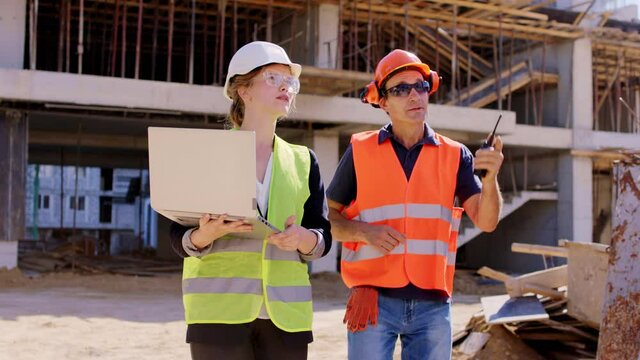 Young engineer lady with a laptop walking beside the foreman middle-aged they analyzing the plan of the construction site wearing safety helmets and goggles