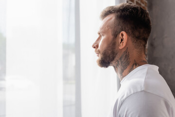 side view of bearded, tattooed man looking away while standing near window at home