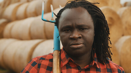 Portrait of young african farmer with pitchfork over his shoulder in front of haystack. High...