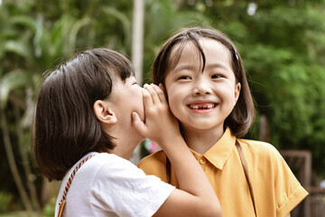 Naklejka premium Two little sister girls whisper in ear at park outdoor.