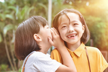 Two little sister girls whisper in ear at park outdoor.
