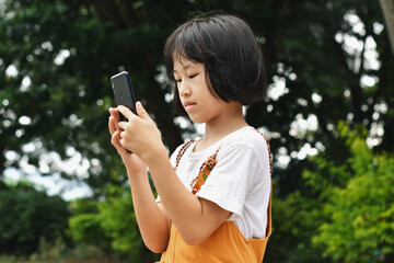 Happy child asian girl using mobile smartphone in the park. Kid is smiling and looking at screen of devices. Concept of communication, technology.