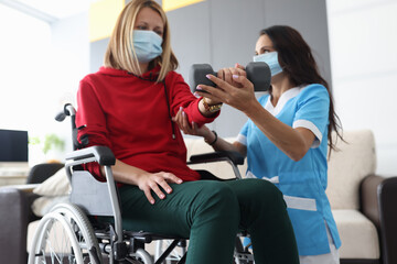 Physiotherapist doctor in medical mask helps to raise dumbbell to disabled woman in wheelchair at...