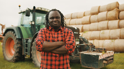 Young african farmer with crossed hands over the chest in front of tractor and haystack. High quality photo