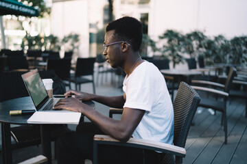 Skilled dark skinned male entrepreneur freelancer in eyewear watching videos and checking mails during remote job on cafe terrace, serious african american developer man typing on laptop computer