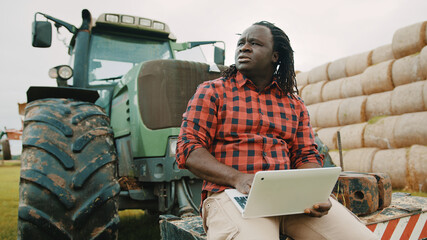 Young african farmer using tablet while sitting on green tractor. Haystack in the background. High...