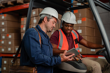 Two male workers wearing hard hats holding digital tablet researching location of parcel while driving factory card in warehouse.