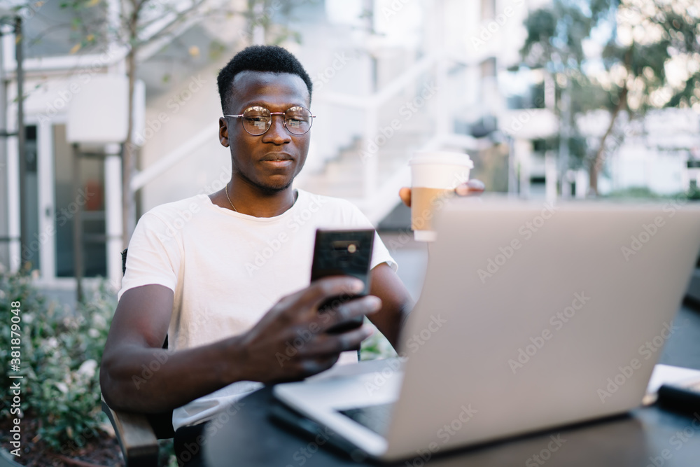 Poster pensive prosperous male freelancer sitting near laptop computer for remote job using smartphone for 
