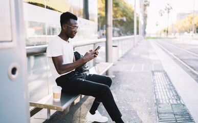 Young dark skinned male traveler in casual wear using app on smartphone for checking public transport schedule, african american man browsing on mobile phone waiting for tram on urban stop
