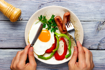 sunny side up. Fried eggs with sausages and vegetables on a light wooden table. Two hands are...