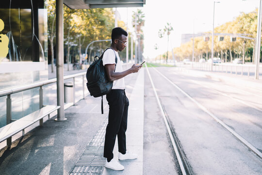 Handsome trendy dressed male tourist checking navigation via application on smartphone with trafic cand public transport schedule. young african american man waiting for bus on station use cellular