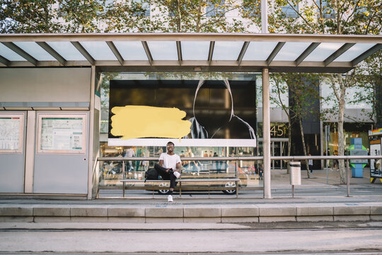 Handsome dark skinned male in casual wear sitting on modern bus stop with advertising banner on urban setting, smiling african american hipster guy waiting for tram satisfied with public transport
