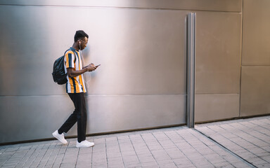 Ethnic student with smartphone walking along light colored wall