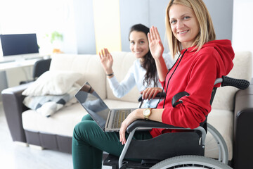 Young disabled woman in wheelchair with helper is sitting by laptop screen and waving hello. Social...