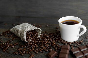 Coffee beans scattered from a linen bag on a wooden table. Cup of coffee and bar of dark chocolate. Mug of black coffee. Morning coffee with a chocolate.