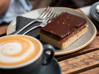 cup of coffee with chocolate brownie on a plate. Brownie in focus, coffee blurry in background
