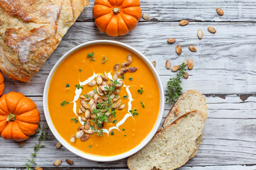 Fresh pumpkin soup garnished with sour cream, toasted pumpkin seeds and thyme with homemade artisan bread over a white wood background. Flatlay.