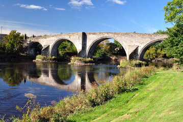 Fototapeta na wymiar View of Ancient Stone Bridge & River with Grassy River Bank against Blue Sky 