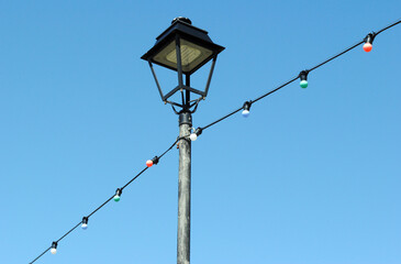 Fototapeta na wymiar Close Up of Pole with Metal Lighting Lantern & Decorative Lights against Blue Sky 