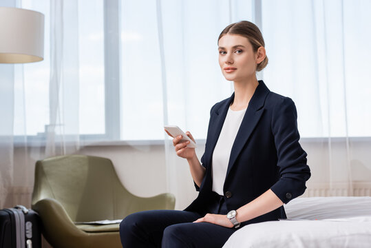 businesswoman in suit holding smartphone and looking at camera while sitting on bed in hotel room