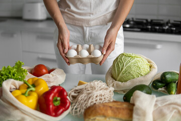 Woman holding eggs tray with a reusable shopping bags with vegetables on a table on a kitchen at home. Zero waste and plastic free concept. Mesh cotton shopper with vegetables. Ecology.