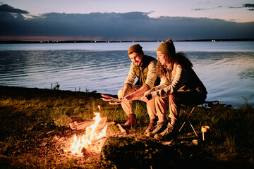 Positive young couple sitting at lake shore and making sausages on fire during night camping