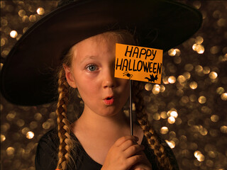 Little pretty girl witch in black wizard hat holds greeting Happy Halloween card and says Boo....
