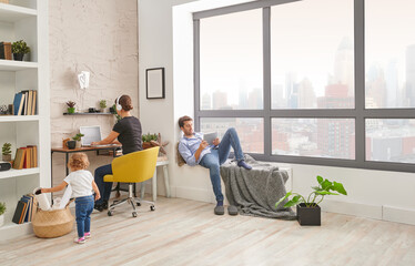 Mom dad and baby in the living room spend time, mother is working and dad is looking tablet.