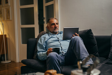 middle aged senior man relaxing on sofa and using tablet computer at home