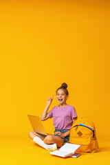 happy schoolgirl with backpack, laptop and books is sitting on yellow background