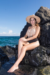 A caucasian woman with sun hat sits in the sun on a coastal rock looking at the camera. Rocky Beach Background