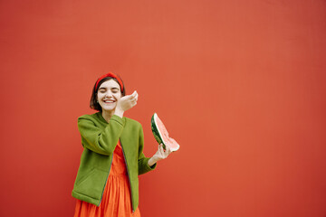 happy brunette girl Bob cut hair style red dress and green jacket holding a piece of watermelon and laughing. Red wall background. Having fun youth concept. hair accessories, mole on the face Teenager