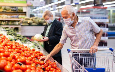 mature man in mask and gloves with covid picks tomatoes in vegetable section of supermarket