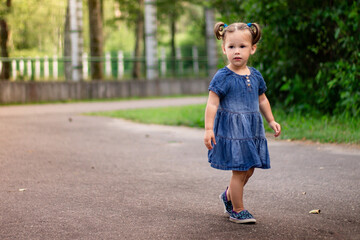 Fototapeta premium Little smiling cute dark-haired girl 1-3 in a denim dress walks along the road in the park on the backdrop of greenery.