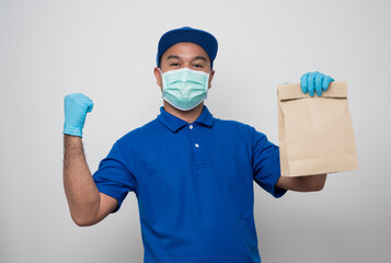 Obraz na płótnie Canvas Happy asian delivery man in blue uniform wearing protection mask and medical rubber gloves giving the paper bag to customer on isolated white background. Safety deliver concept..