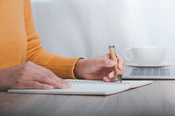 Cropped image of business woman sitting at table and taking notes in notebook, On table is laptop and cup of coffee.