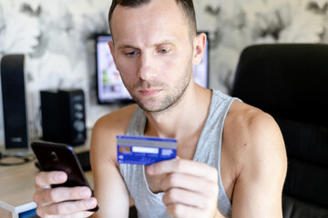 serious man sitting at a computer table with a credit card and a smartphone in his hands