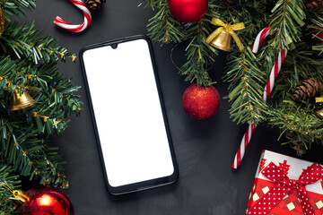 A smartphone with a white screen lies on a dark background covered with Christmas tree branches and Christmas toys.