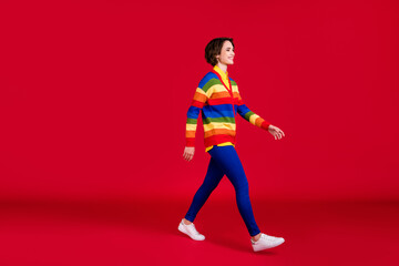 Fototapeta na wymiar Full size profile photo of beautiful pretty young lady good mood walking down street friends meeting wear rainbow jumper cardigan trousers shoes isolated bright red color background