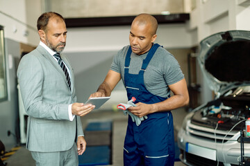 Black mechanic and his manager working on touchpad in auto repair shop. - obrazy, fototapety, plakaty