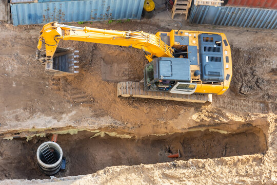 Excavator at a construction site while digging trenches for calcining sewer and drainpipes with a raised bucket, top aerial view.