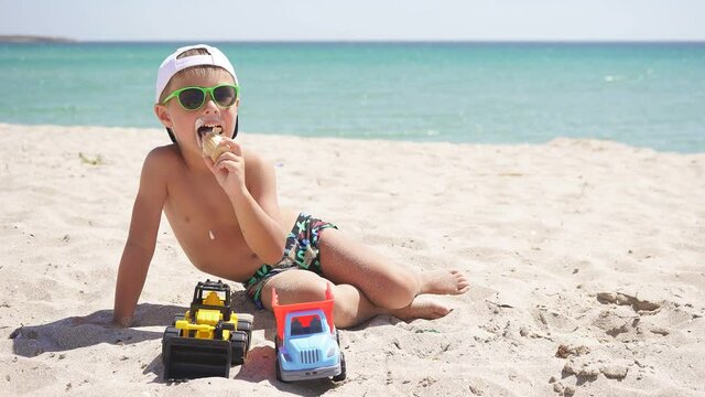 tanned boy wearing a baseball cap eats ice cream while sitting on a sandy beach, against a beautiful azure sea and blue sky on a Sunny day.
