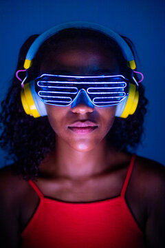 Young woman wearing headphones and futuristic led glasses on blue background - Isolated black woman wearing 3d smart glasses and headphones