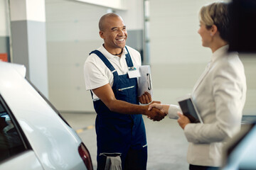 Happy African American mechanic handshaking with businesswoman in auto repair shop.