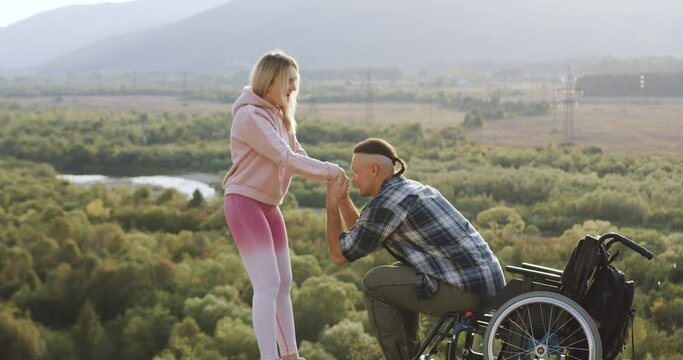 Portrait of good-looking smiling happy couple where disabled man getting up from wheelchair with his wife's help and doing his first steps after desease or accident on the hill on the picturesque